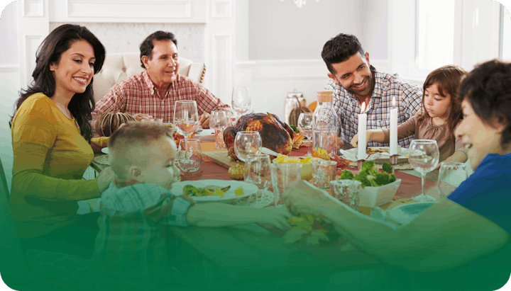 A family gathered around a table enjoying a meal with a roast turkey centerpiece, surrounded by smiling faces, candles, and dishes.