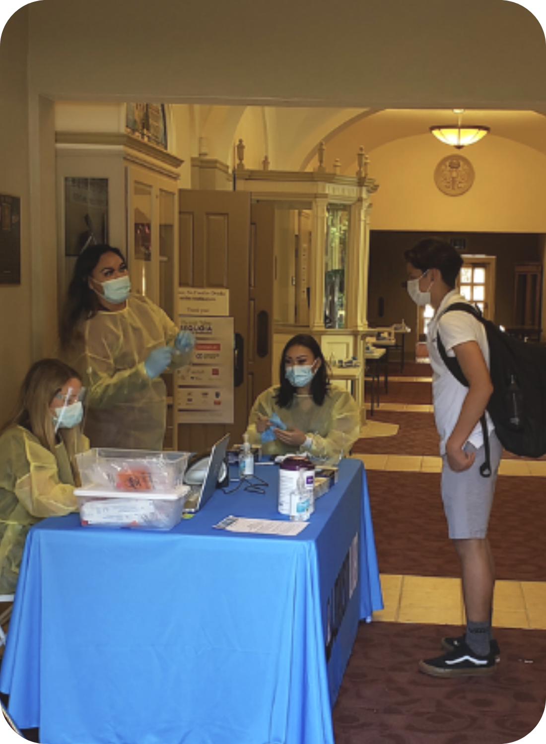 A masked student at a check-in table with healthcare workers in PPE at a vaccination or testing site.