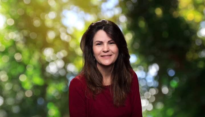 A person with long dark hair and a red top smiles at the camera, with a blurred nature background.