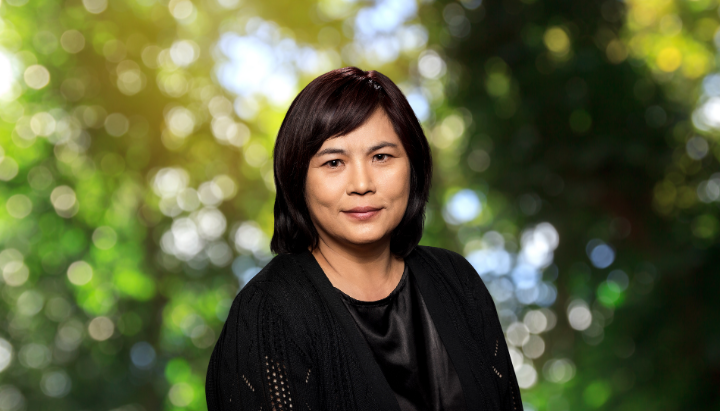 A person in a dark outfit stands against a blurred, leafy background.