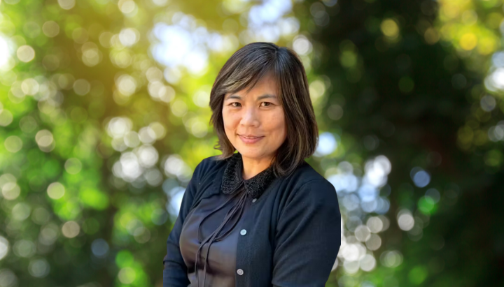 A person in a dark outfit is standing outdoors with a blurred leafy background and bokeh effect.