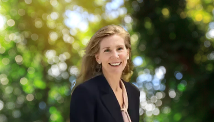 A person smiling, wearing a dark blazer, with a leafy, sunlit background.
