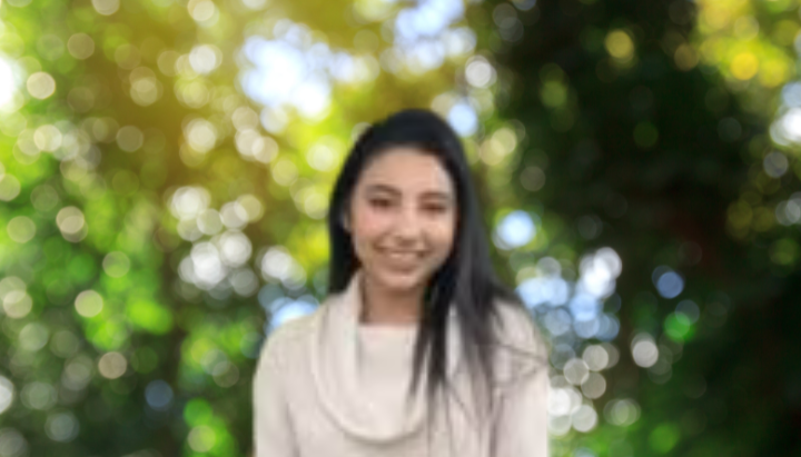 A person with long dark hair is smiling, standing outdoors with a blurred background of green foliage and sunlight.