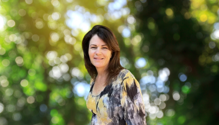A person smiling outdoors, with a bokeh effect and green foliage in the background.