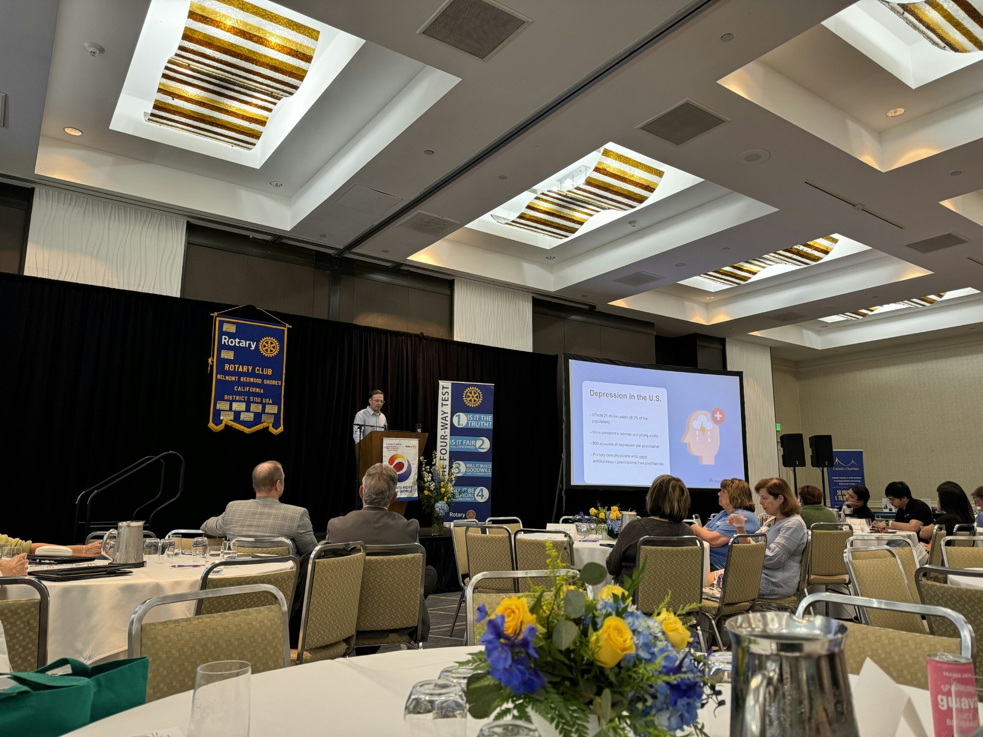 A conference room with attendees and a speaker presenting on "Depression in the U.S." at a Rotary Club event.