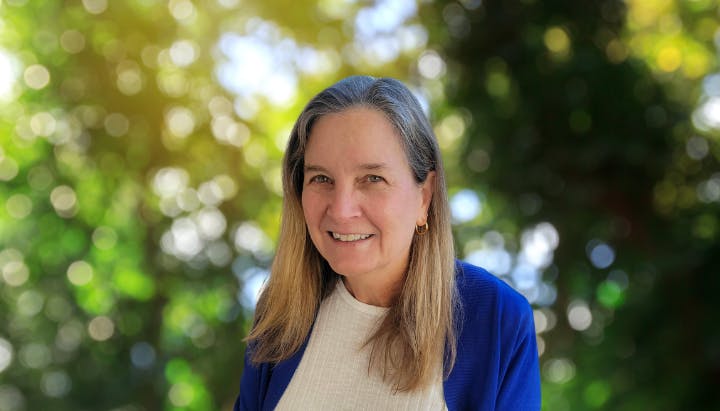 A woman in a blue sweater smiles outdoors with a blurred background of green foliage and sunlit bokeh.