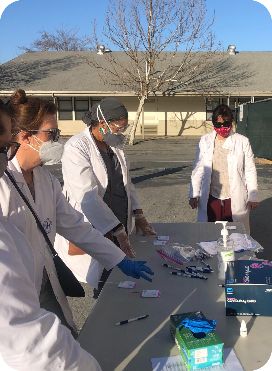 People in masks and lab coats work at a table with COVID-19 test kits and supplies.