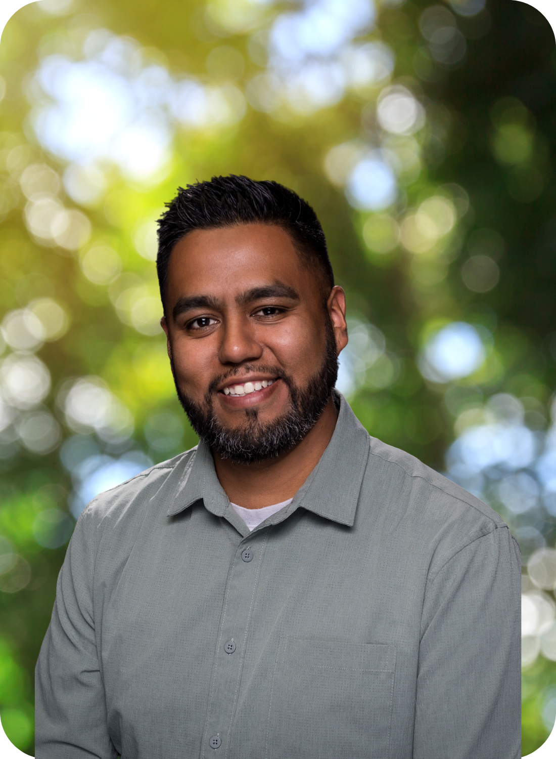 A man in a gray shirt stands against a blurred, sunny, green background, smiling at the camera.