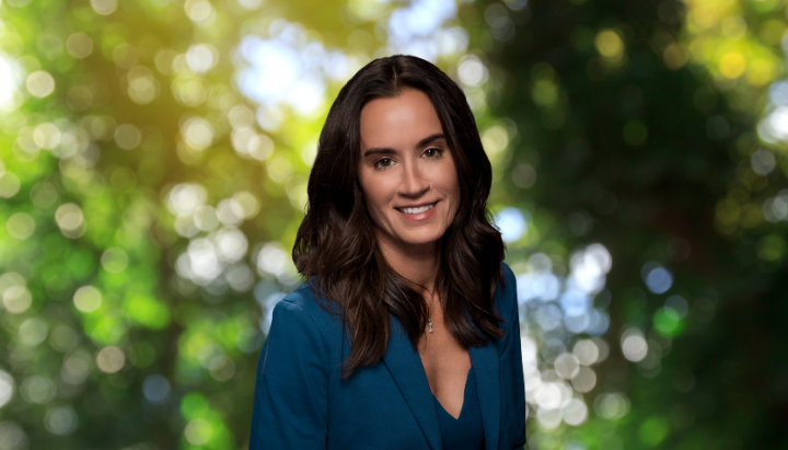 A woman in a blue blazer stands in front of a blurred green and yellow nature background, smiling.
