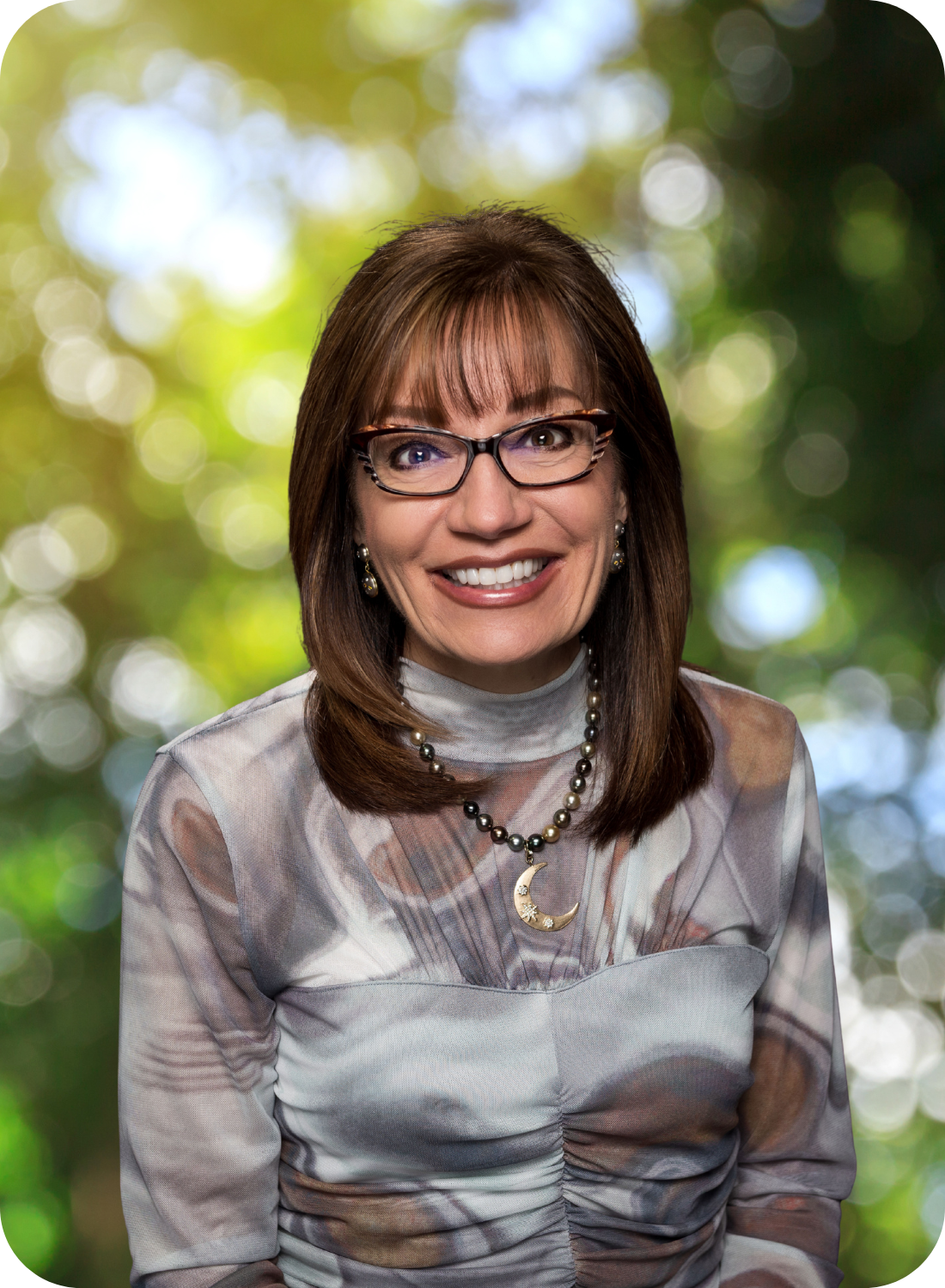 A person with glasses, wearing a patterned shirt and a necklace with a crescent moon pendant, smiles against a blurred green background.