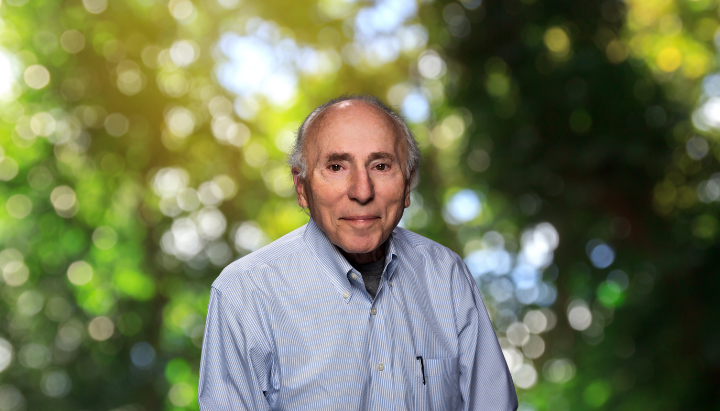An older man in a light blue shirt standing in front of a blurred natural background with green foliage.
