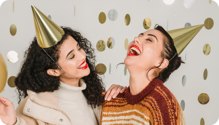 Two smiling women in party hats celebrate joyfully against a backdrop of golden dots, radiating happiness and friendship.
