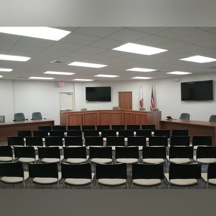 A meeting room with rows of chairs facing a podium, two flags, and screens on the wall.