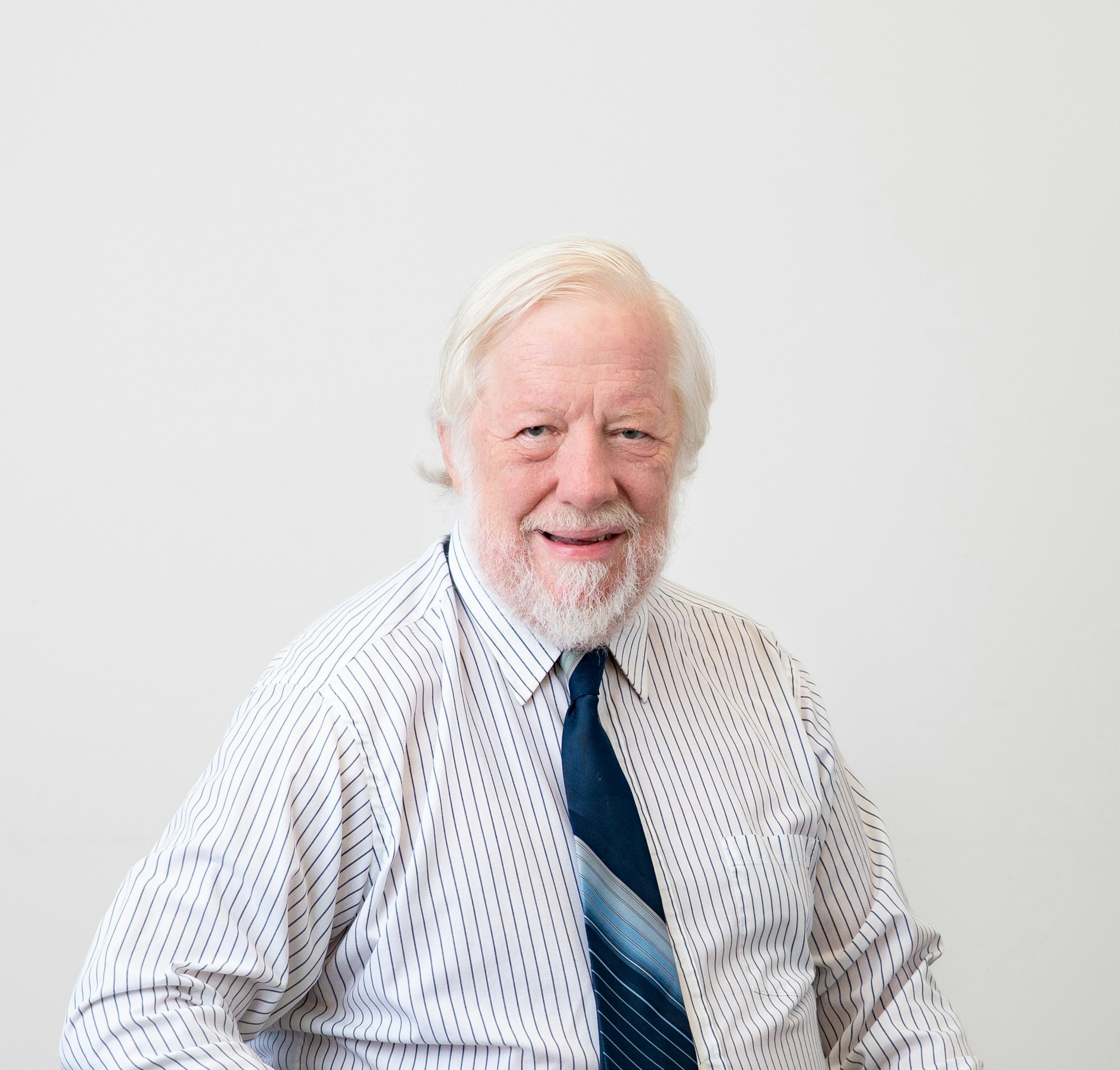 An older man with white hair and beard wearing a striped shirt and blue tie, smiling against a plain background.