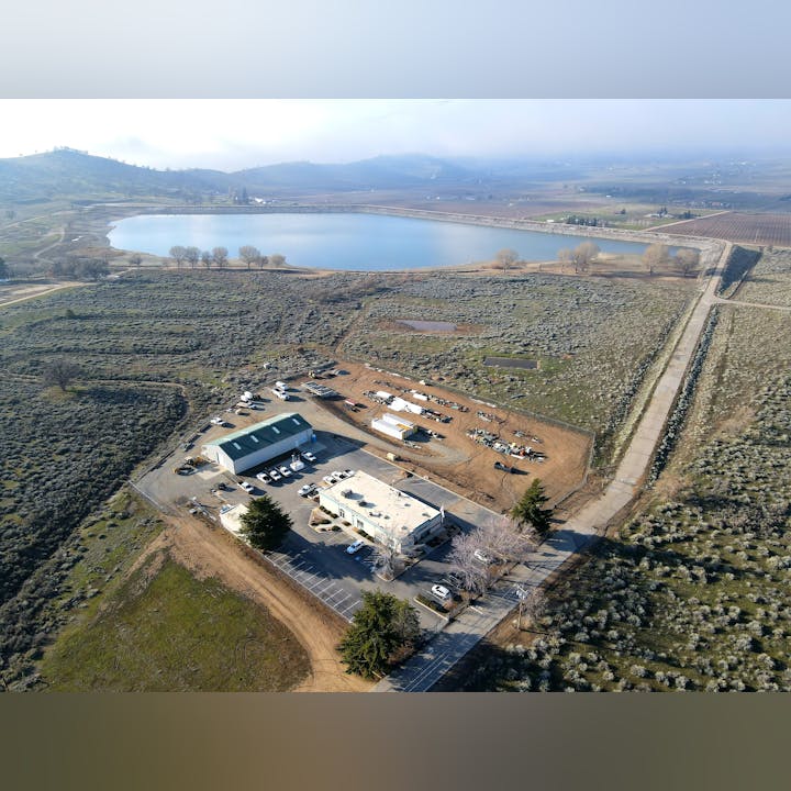 Aerial view of a rural facility with buildings, parked vehicles, and a large body of water near fields and hills.