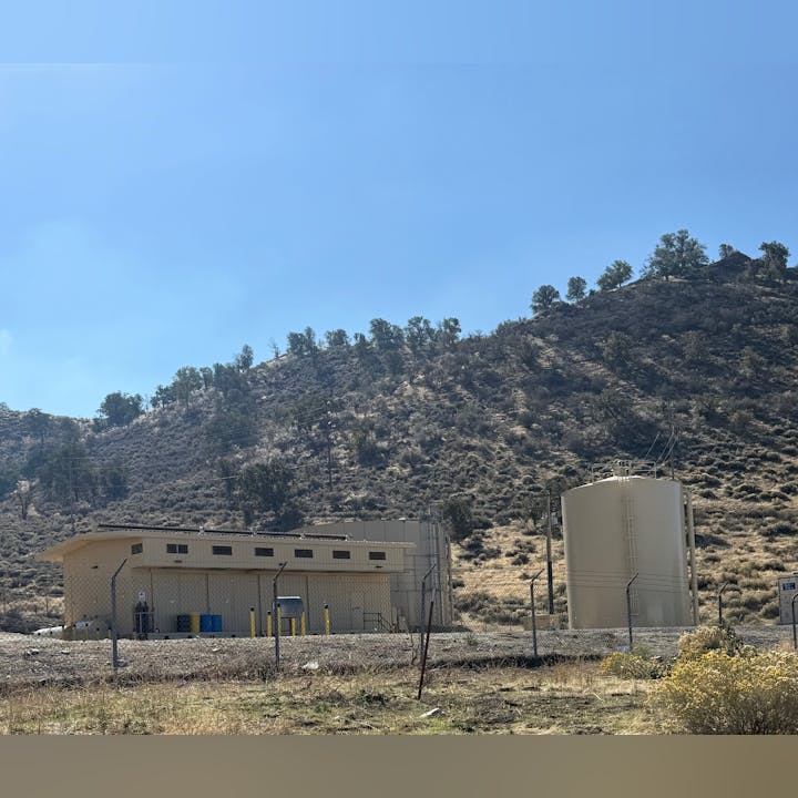 A fenced industrial facility with a building and a large tank, set against a hillside with sparse trees and under a clear blue sky.