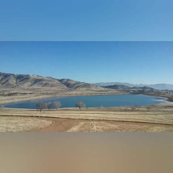 A scenic landscape with a blue lake, dry fields, scattered trees, and hills under a clear blue sky.