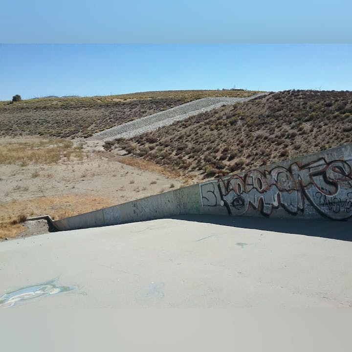A concrete slope with graffiti, adjacent to dry, rocky terrain under a clear blue sky.