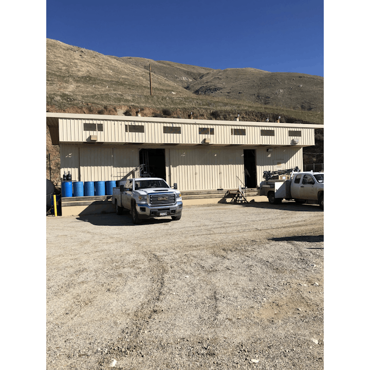 A building with parked trucks outside, located on a dirt area with grassy hills and a clear blue sky in the background.
