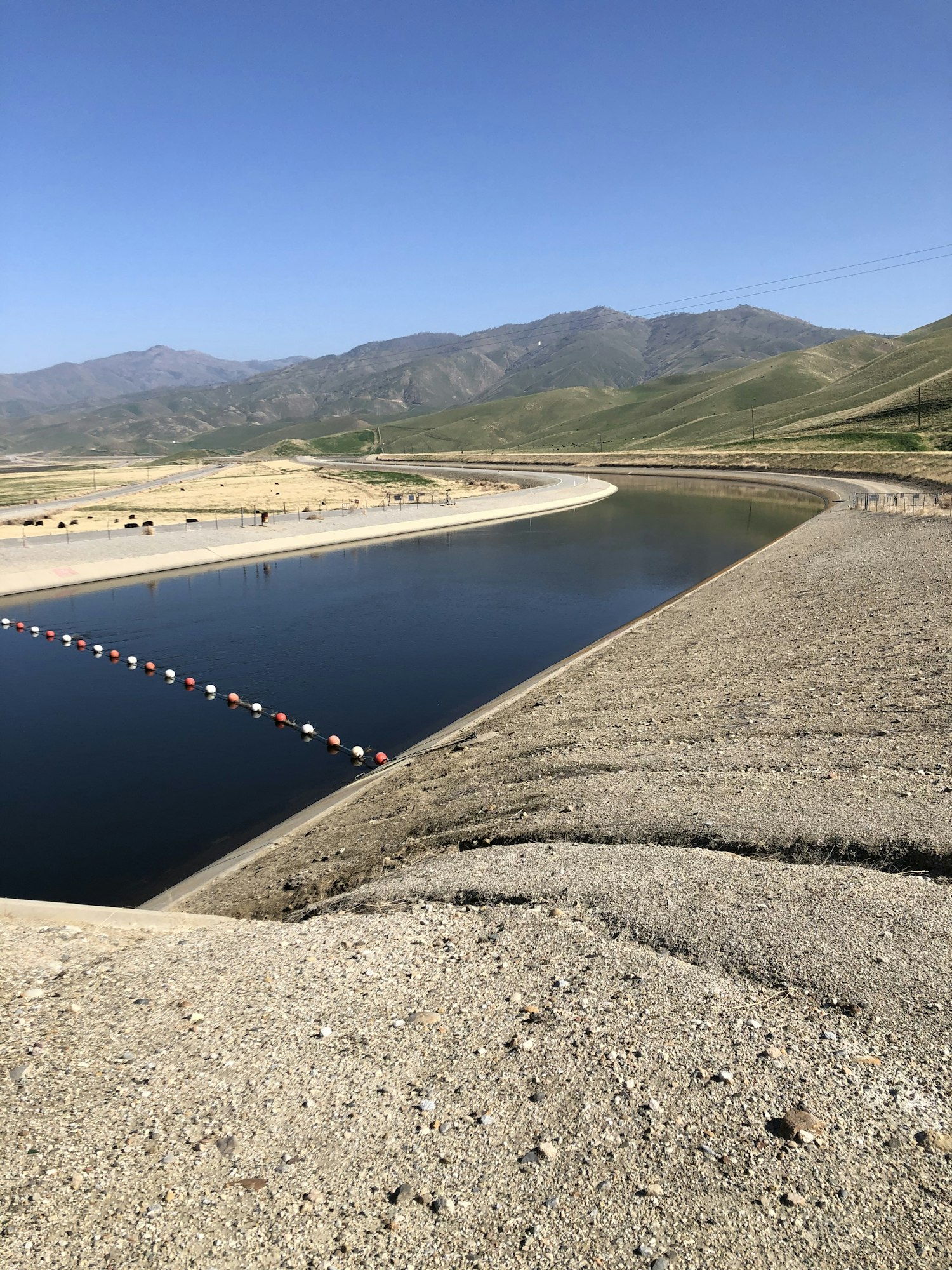 A canal with concrete banks and a line of buoys, surrounded by hilly landscape and a clear blue sky.