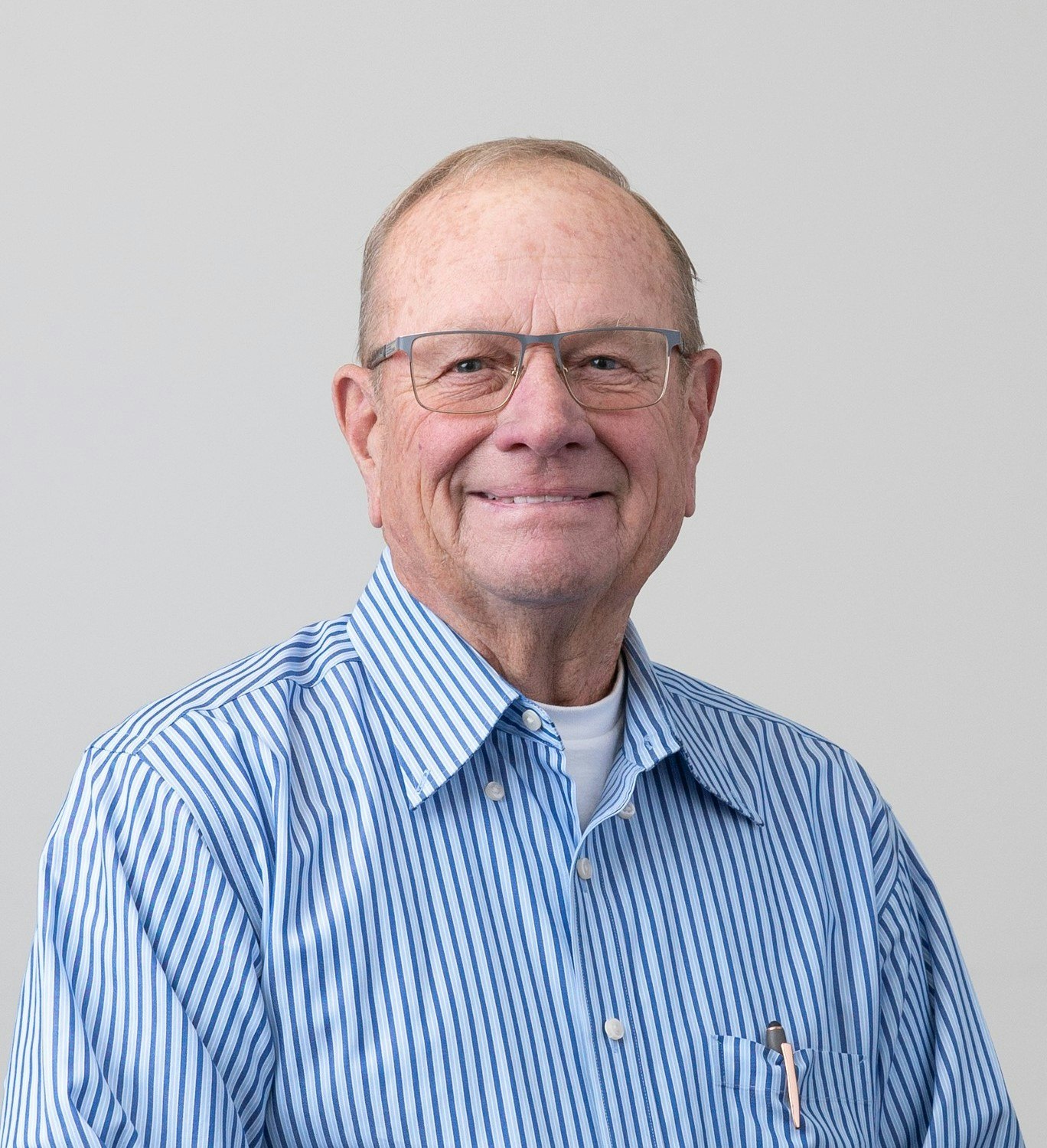 An older man wearing glasses and a blue striped shirt, smiling against a plain background.