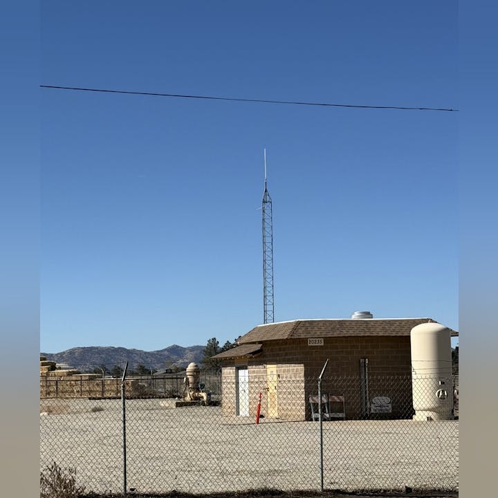 A fenced utility area with a building, tower, and tanks under a clear blue sky.