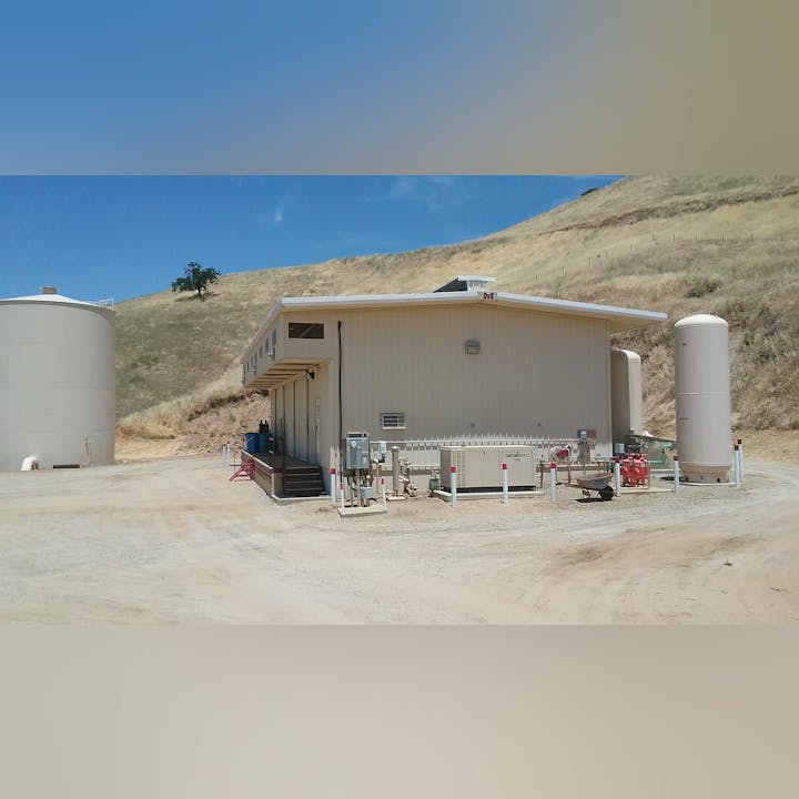 Industrial facility with large storage tanks, a building, and equipment on a dirt road in a hilly area.