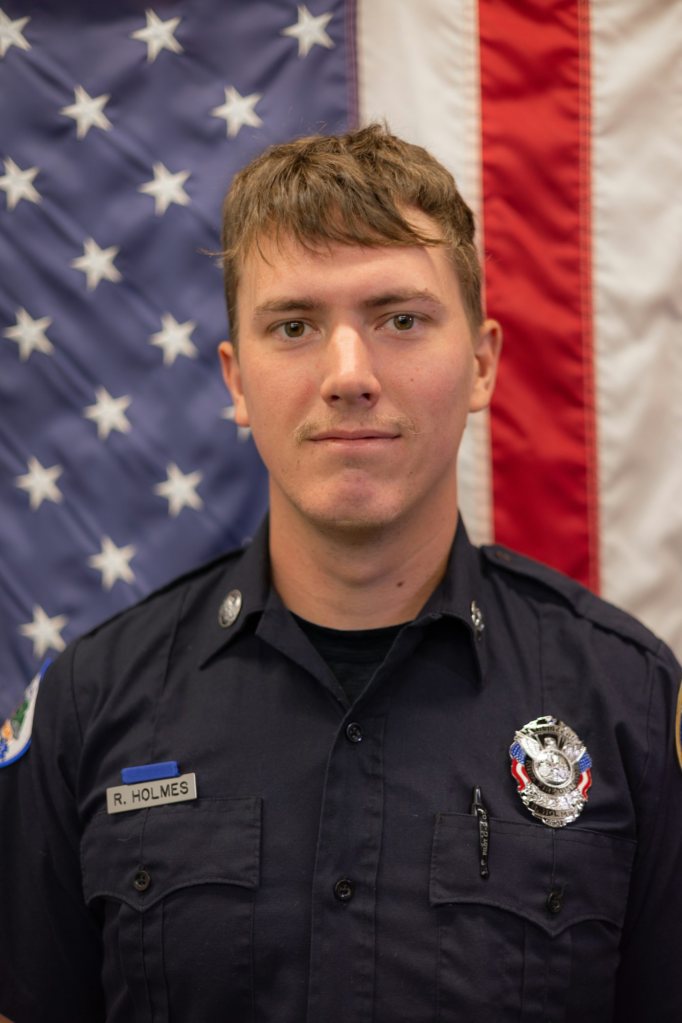 A person in a uniform with a badge, in front of an American flag.