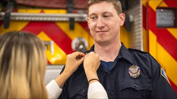 A woman is pinning a badge on a smiling man in uniform, with a fire truck in the background.