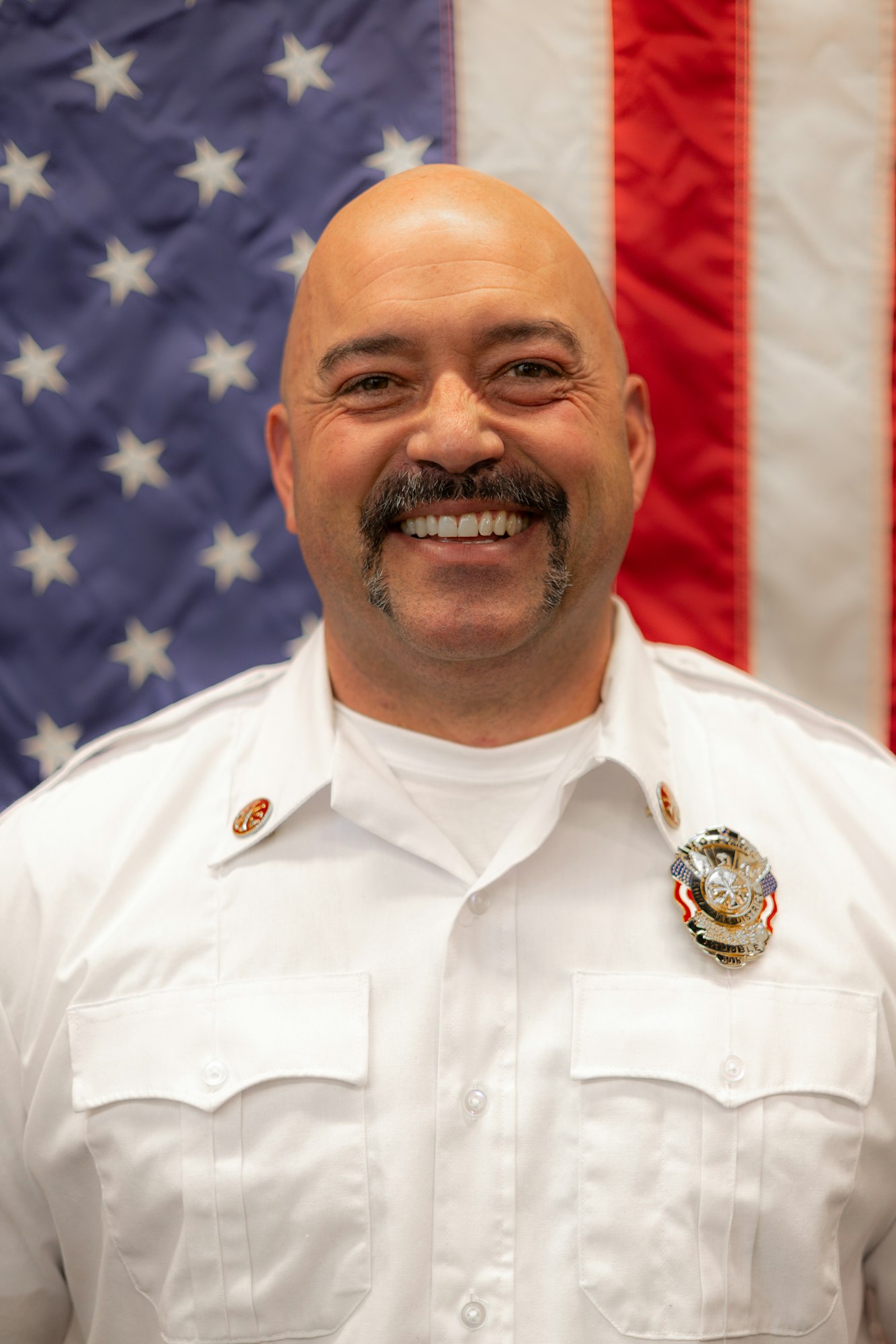 A smiling man in a white uniform with a badge in front of the American flag.