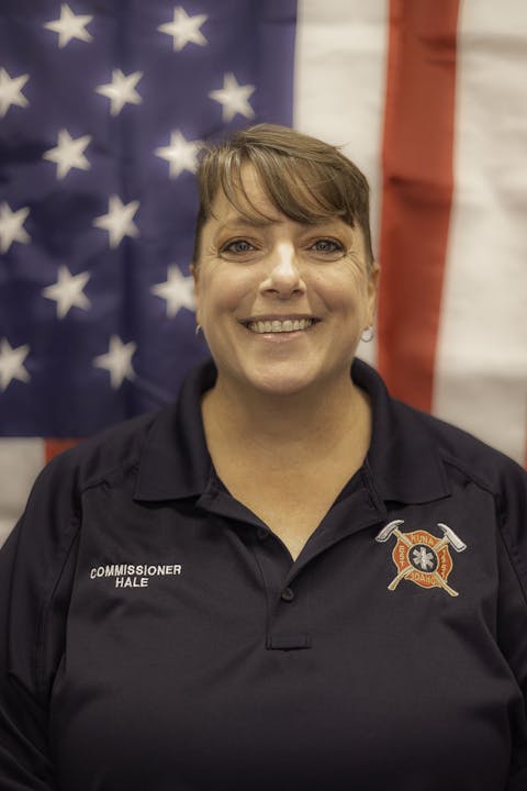A smiling woman in a uniform with "Commissioner Hale" on it, standing in front of an American flag backdrop.