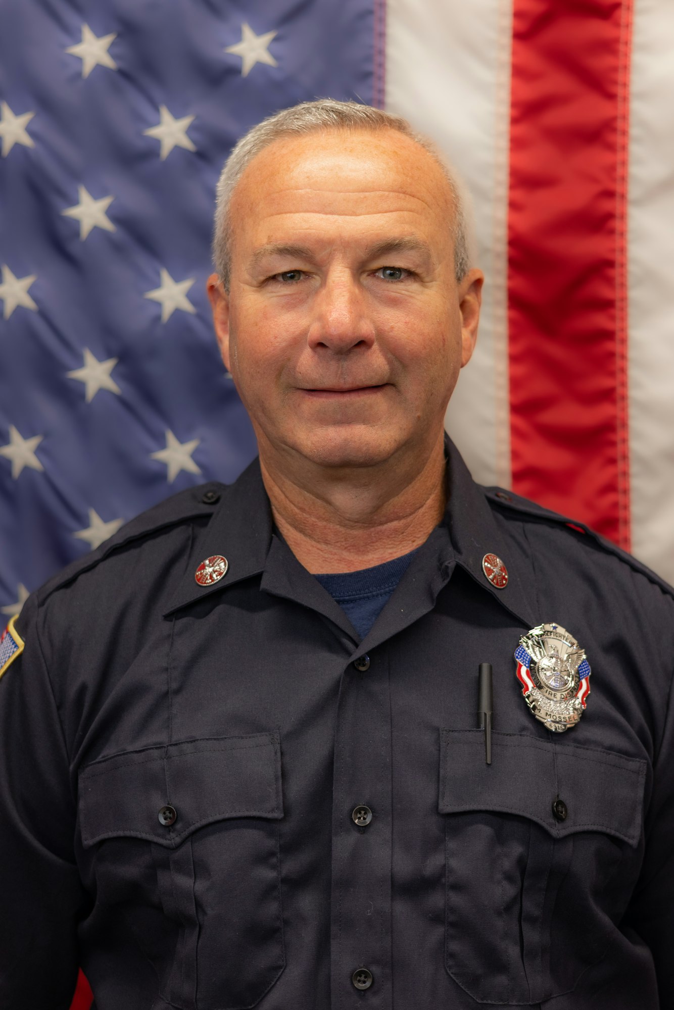 A firefighter in uniform stands in front of an American flag backdrop.