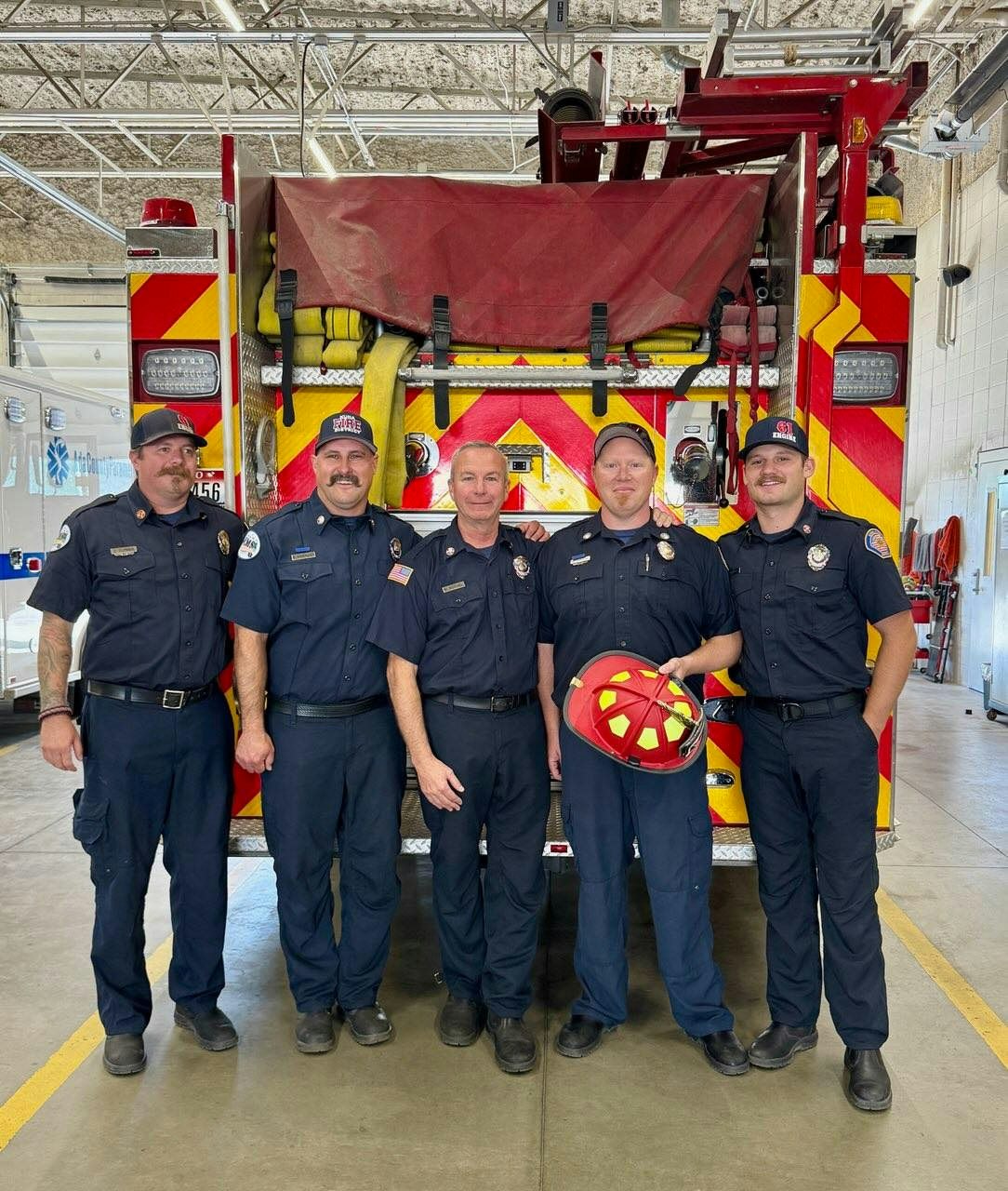 Five firefighters in uniform stand in front of a fire truck in a garage.