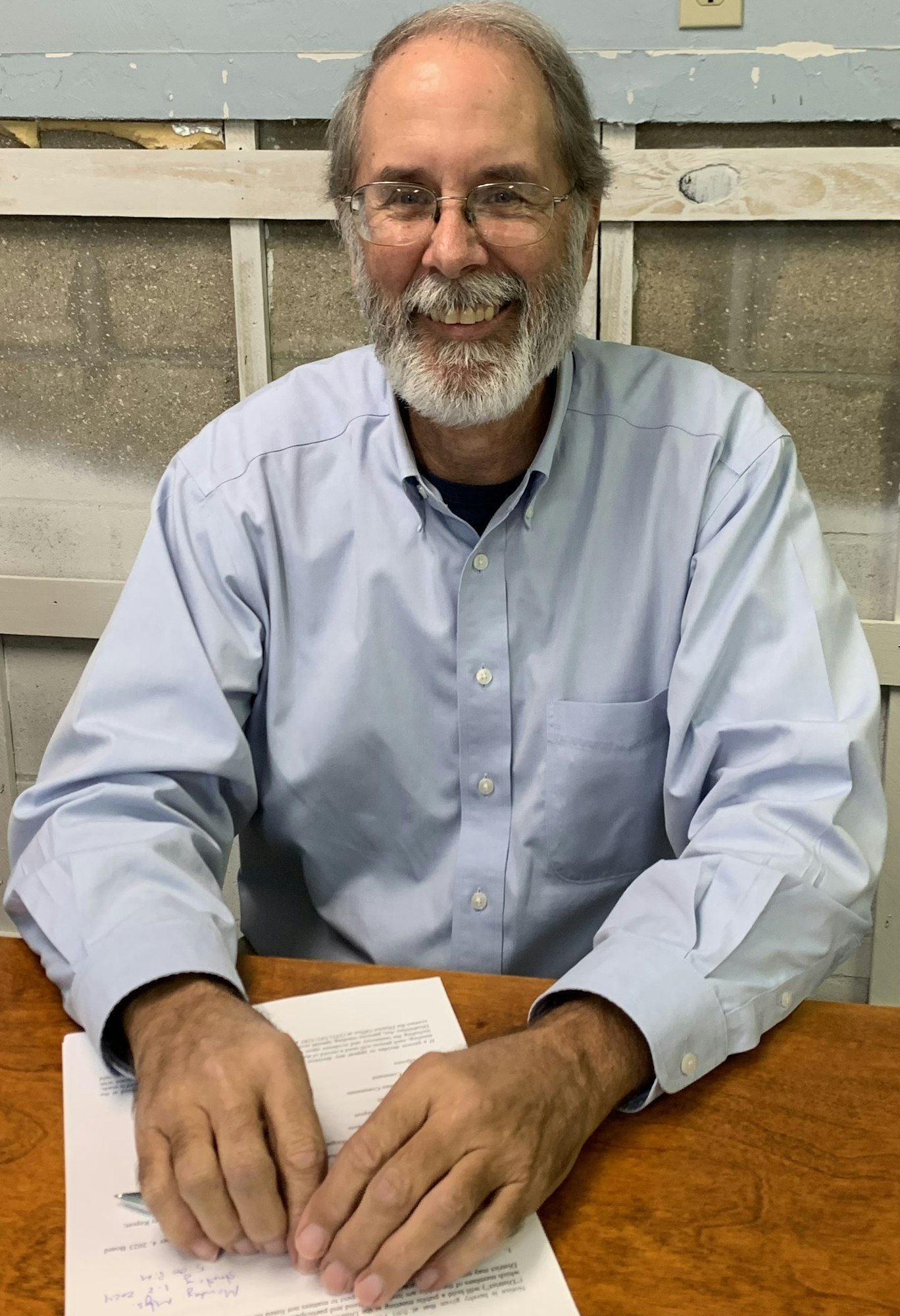 A smiling man with glasses and a beard sits at a table, hands on documents, in a casual setting.