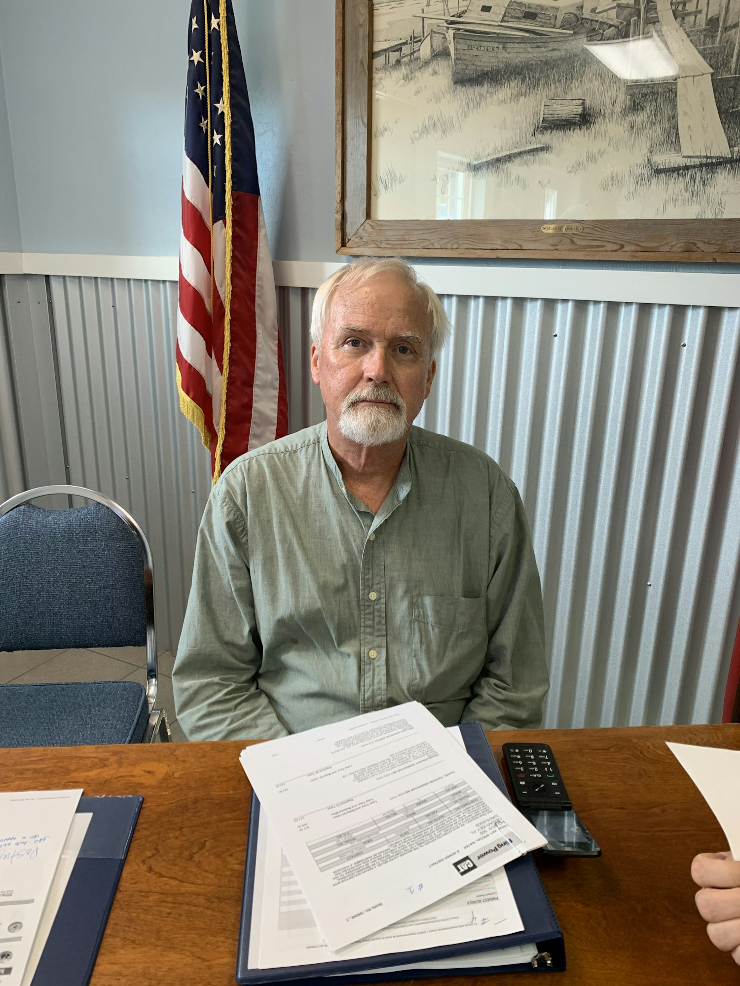 A man with a beard sits at a table with paperwork, an American flag in the background, and a phone nearby.