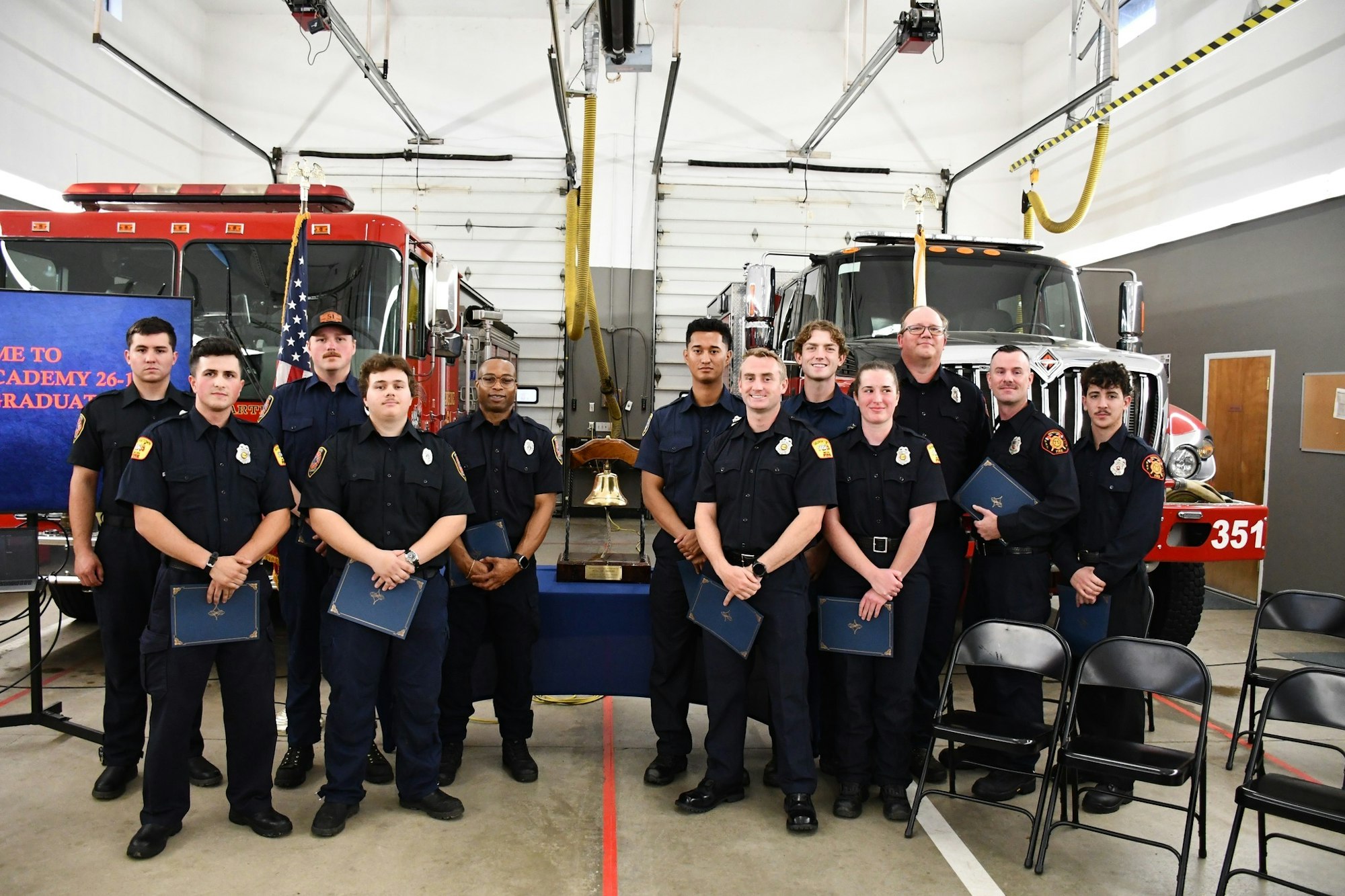 A group of fire academy graduates poses in front of fire trucks, holding diplomas and celebrating their completion of training.