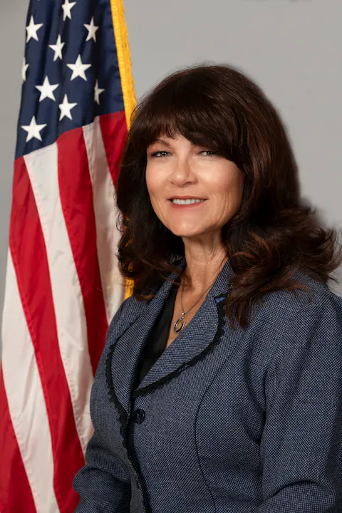The image shows a woman smiling in front of an American flag, wearing a stylish blazer.