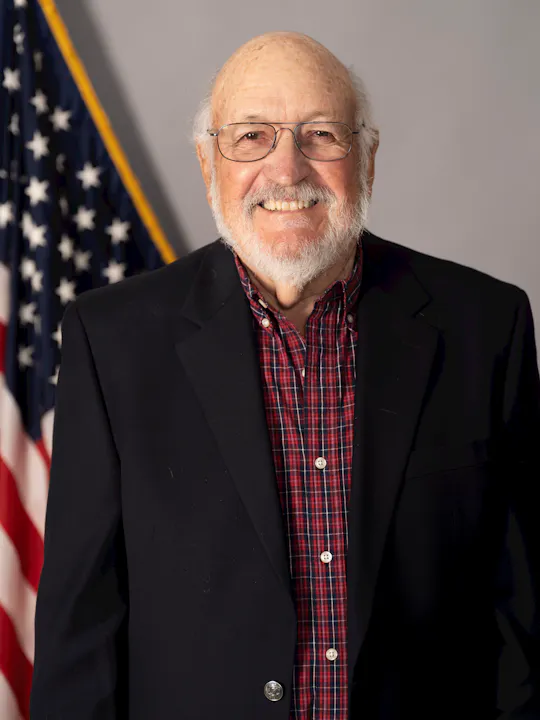 A smiling elderly man with glasses, wearing a black blazer over a plaid shirt, stands in front of an American flag backdrop.