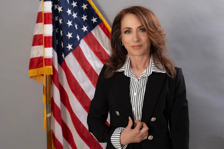 A woman in a black suit with a striped shirt stands next to the American flag, looking confidently at the camera.