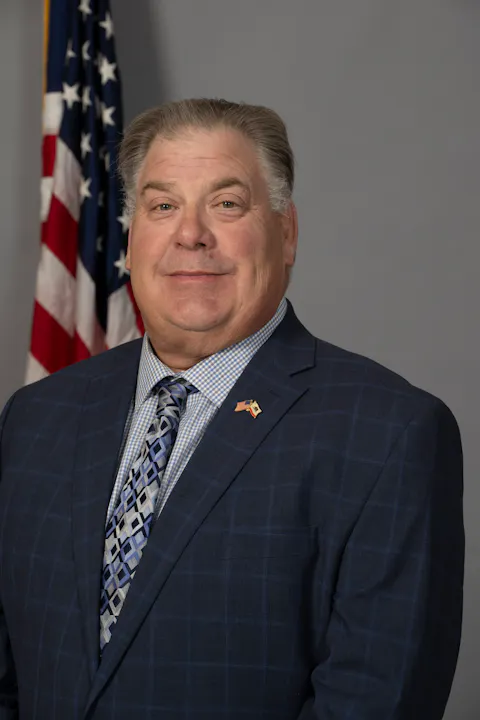 A man in a blue plaid suit and patterned tie poses in front of an American flag against a gray background.
