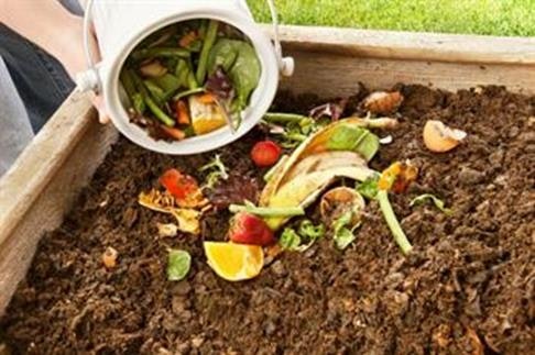 A person is dumping food scraps and organic waste into a compost bin filled with dark, rich soil.