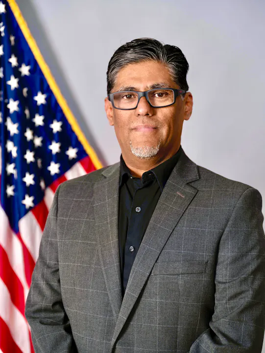 A man in a suit stands in front of the American flag, posing confidently for a formal portrait.