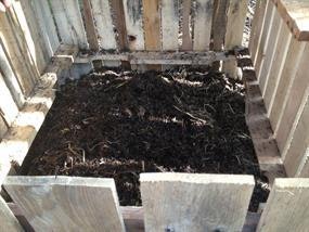 The image shows a compost bin made of wooden pallets, filled with dark, nutrient-rich compost.