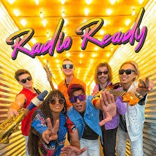 A lively group of musicians poses in colorful outfits with instruments, set against a bright, illuminated backdrop.