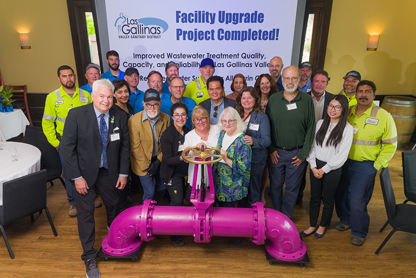 Group of people celebrating a facility upgrade with a banner and a large valve in the foreground.