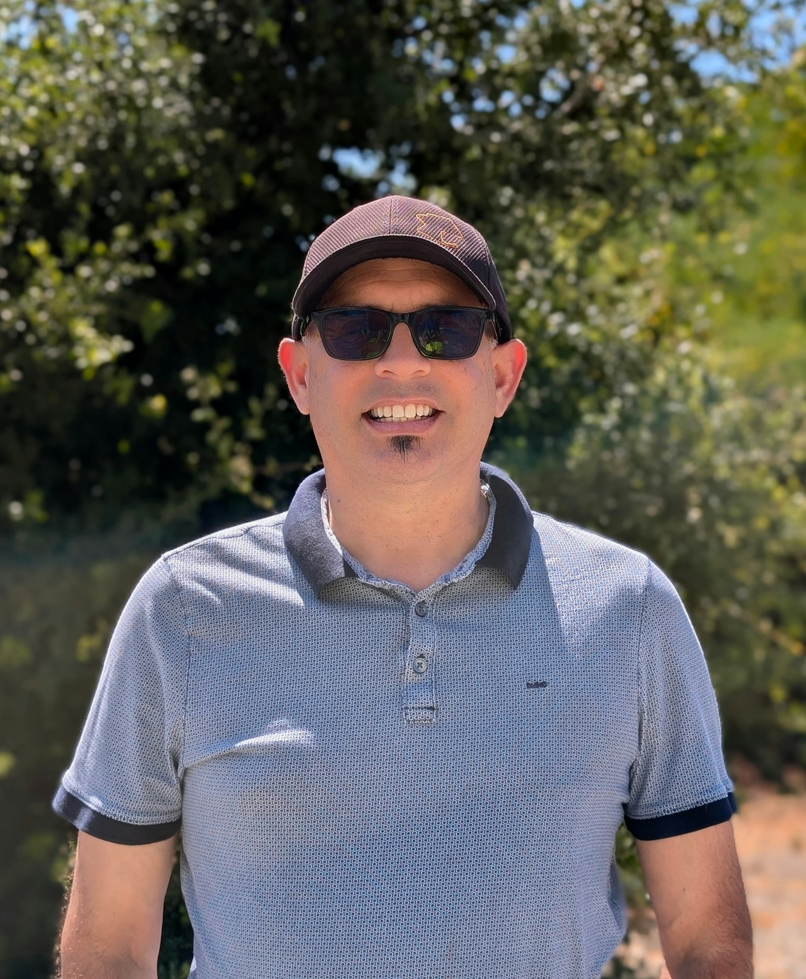 A person wearing sunglasses and a cap, smiling outdoors with trees in the background.