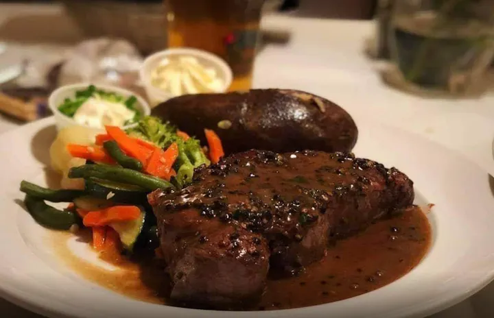 A plated meal featuring a steak with sauce, mixed vegetables, and a baked potato, alongside a drink.