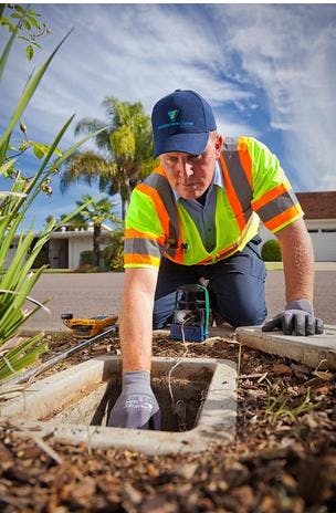 A worker in a reflective vest and cap checks an underground utility box near a suburban street, with tools nearby.