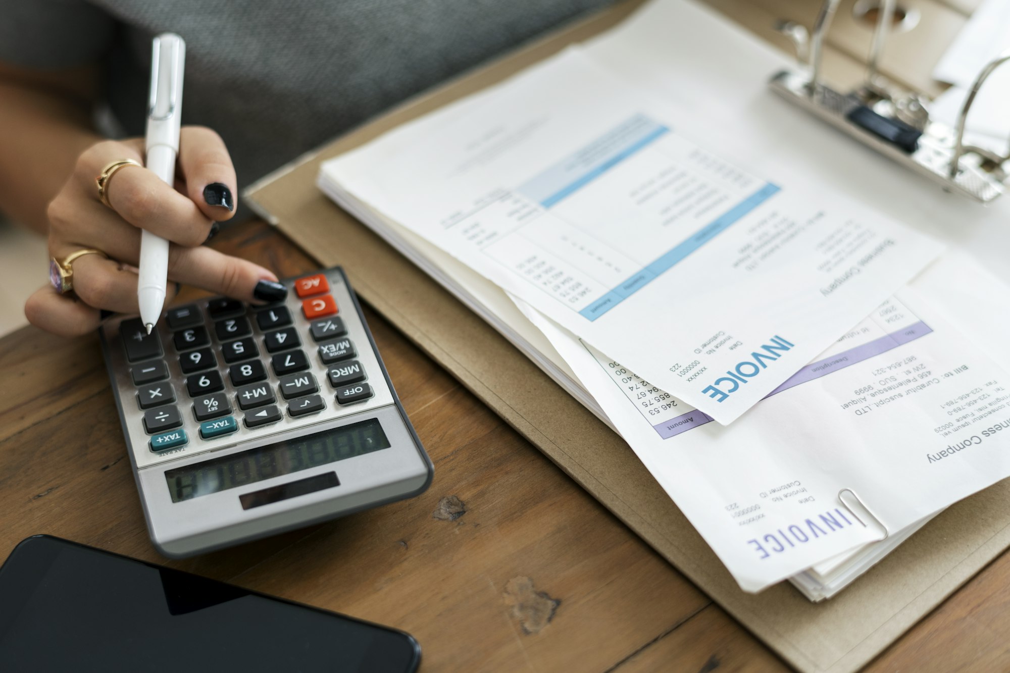 An individual calculates figures with a calculator, surrounded by invoices and papers on a wooden desk.