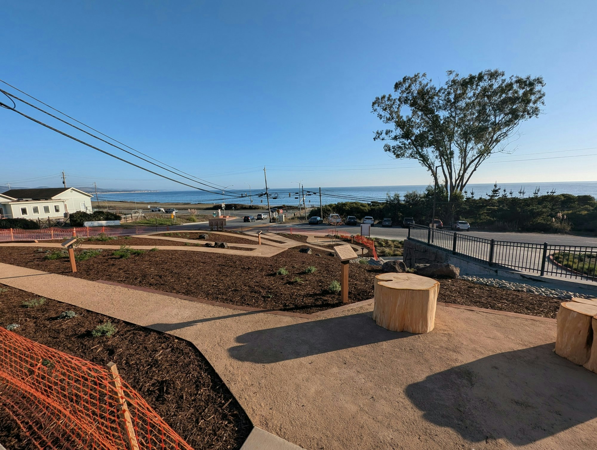 A seaside park view with dirt paths, wooden benches, and trees, overlooking a road, beach, and ocean under a clear blue sky.